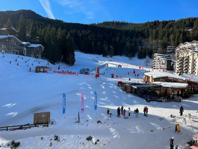 A snowy landscape reveals a busy ski area with people enjoying winter activities. Colorful banners flank the area, while wooden structures, including a bar, blend into the surroundings. The backdrop features a mix of trees and mountain slopes under a clear blue sky.