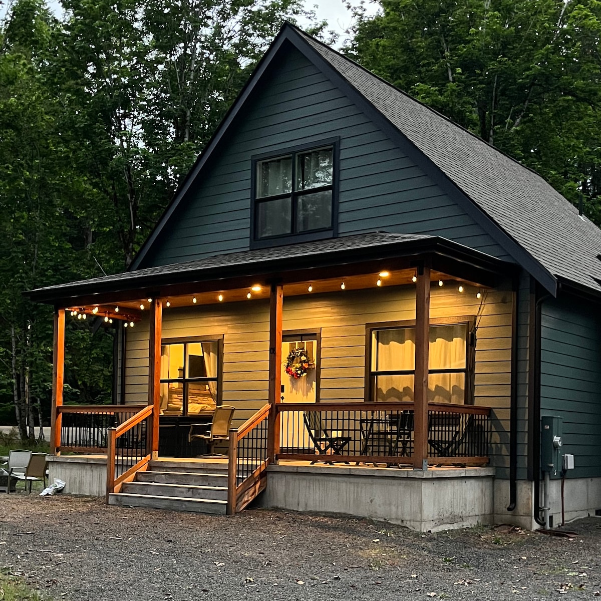 A modern, two-story house showcases a welcoming front porch with string lights. Large windows provide natural light, while a wreath adorns the door. The structure is surrounded by greenery, creating a tranquil atmosphere.