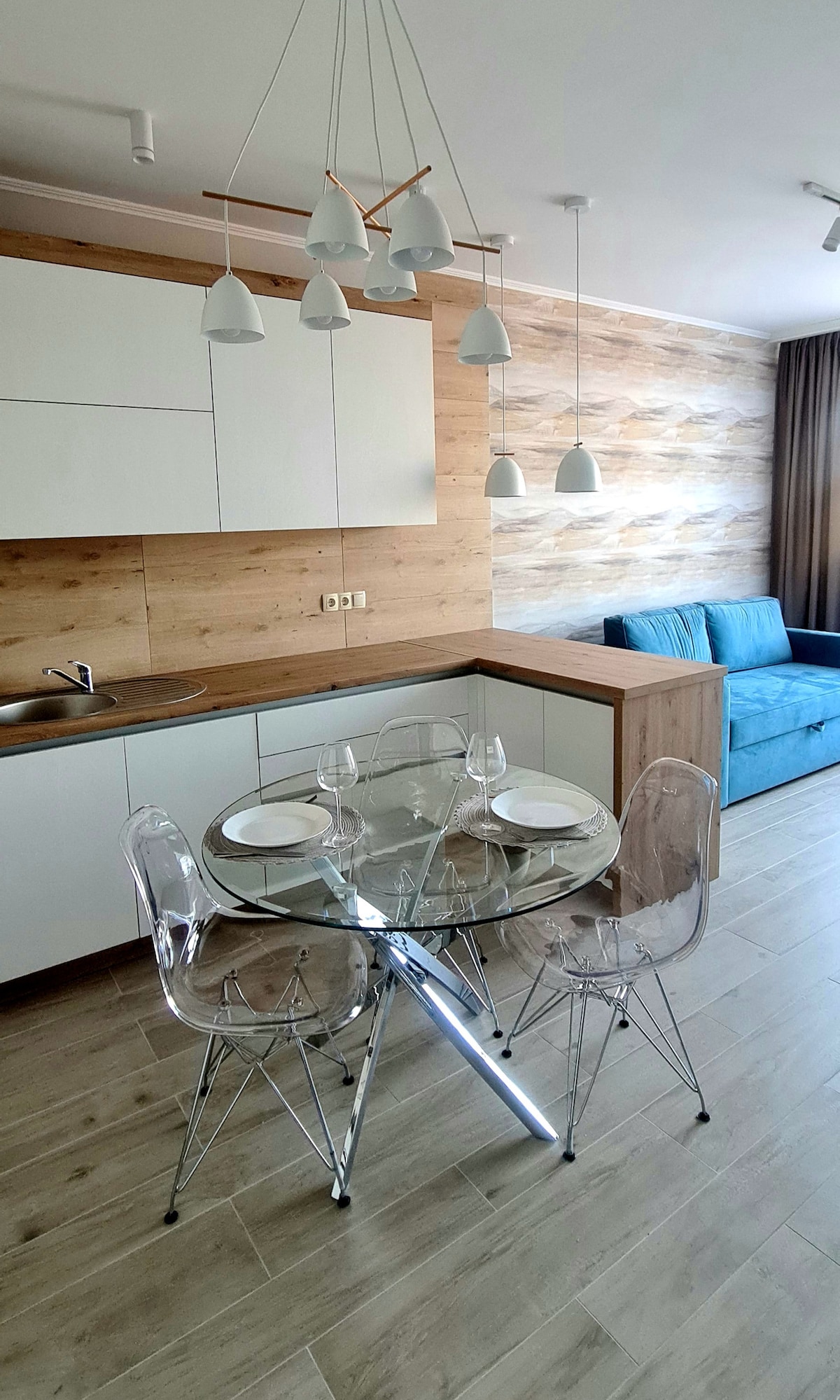 A modern kitchen area is featured, with a circular glass dining table set for four. Transparent chairs surround the table, while a sleek blue sofa is positioned in the background. Soft lighting is provided by hanging pendant lamps above the table.