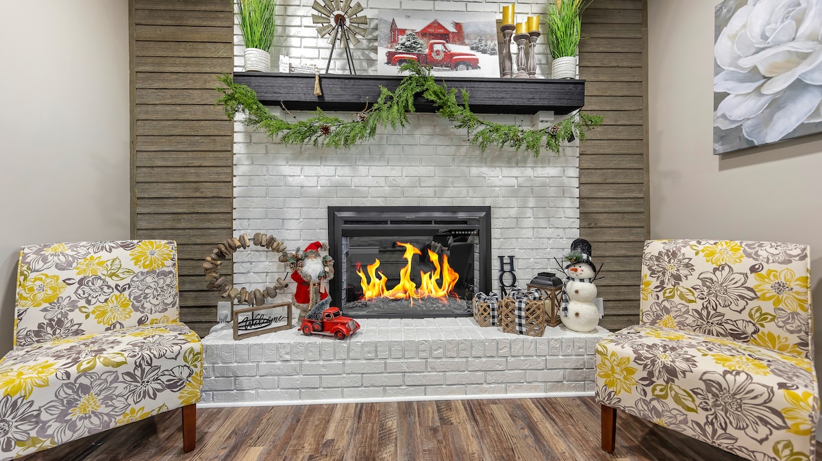 A cozy fireplace is framed by a white brick wall, with a black mantel adorned with decorative items. Two patterned chairs with floral designs are positioned on either side, offering a comfortable seating area. The fireplace features flickering flames, contributing to a warm atmosphere.