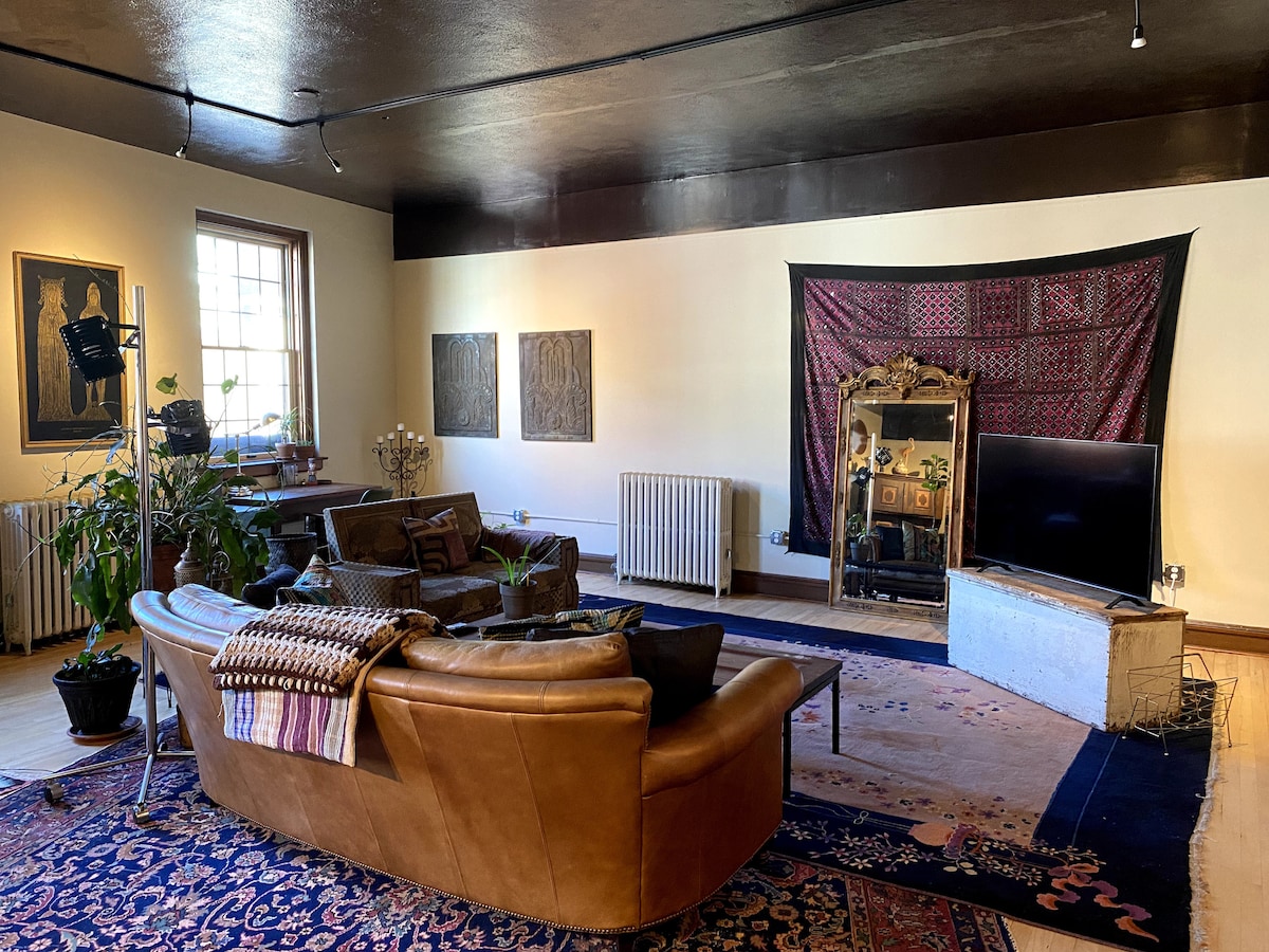 A spacious living area features a large, brown leather sofa positioned around a central area rug. Plants are placed throughout, with a decorative mirror and artwork on the walls. A television is situated on a low table, enhancing the cozy atmosphere.