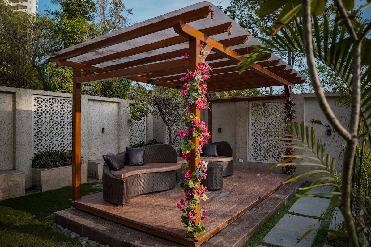 An outdoor seating area is framed by a wooden pergola adorned with blooming flowers. Comfortable furniture provides a relaxing space, while the surrounding greenery adds a touch of nature. The patio floor is designed with tiles, and the ambiance is serene.
