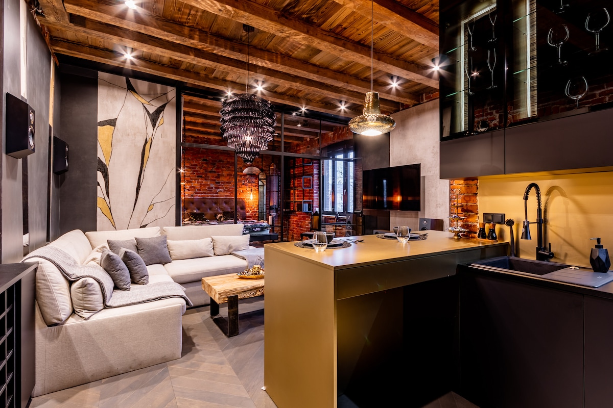 A spacious living area features a comfortable sectional sofa in neutral tones, contrasting with the warm wood beams and exposed brick walls. An elegant light fixture hangs above a sleek kitchen island, which is equipped with modern appliances, creating a stylish and functional space.
