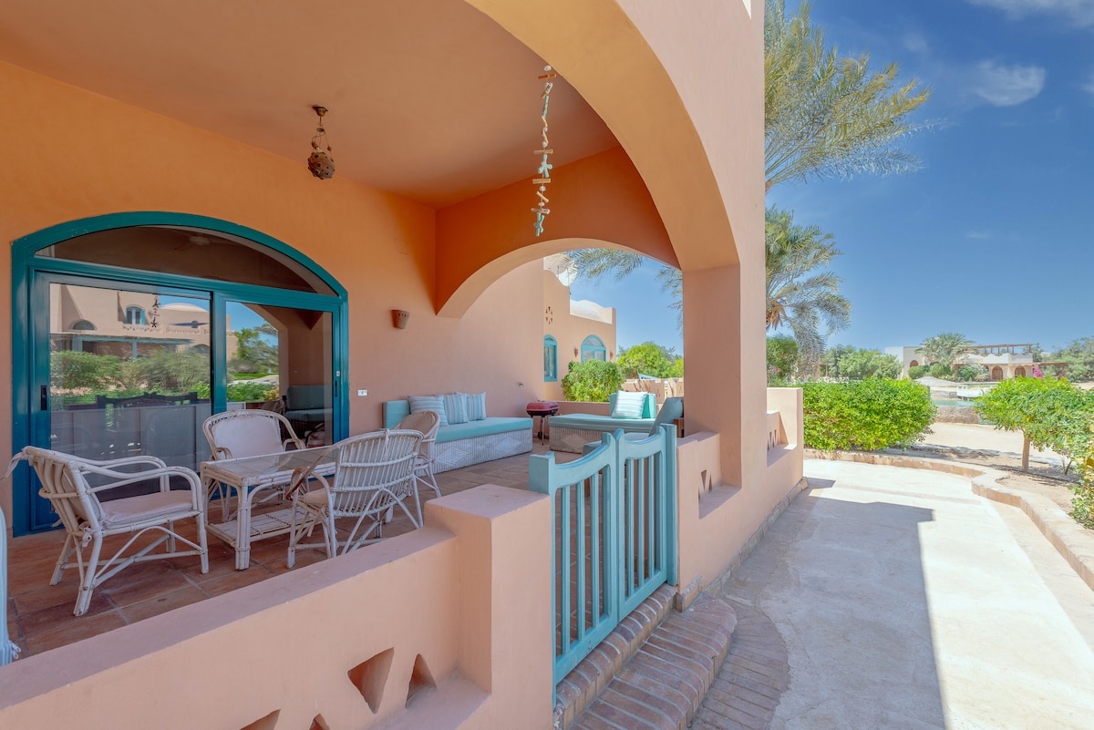 A spacious terrace features multiple seating arrangements including a small table and chairs, surrounded by lush greenery. The entryway showcases arched doorways and vibrant blue accents, inviting open-air relaxation. Sunlight casts soft shadows, enhancing the inviting atmosphere of the exterior space.