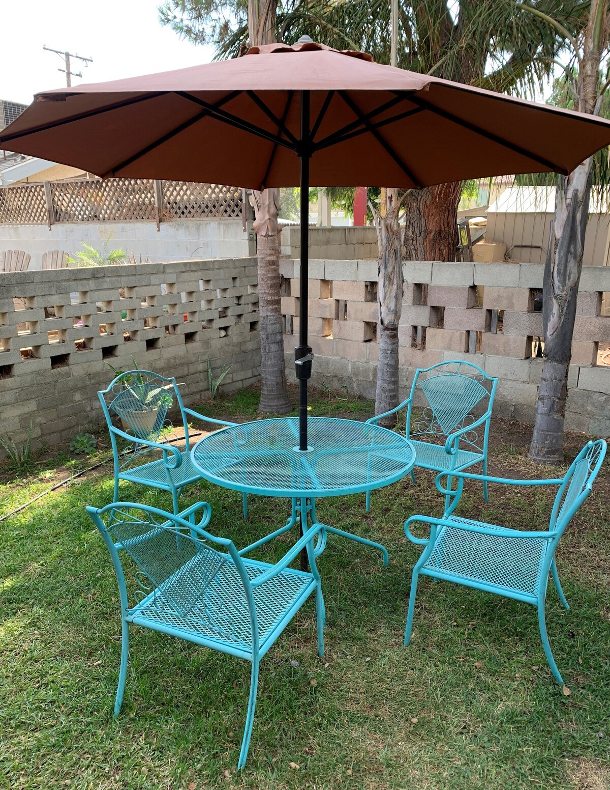 A private garden area is depicted, featuring a round table surrounded by four turquoise metal chairs. A large umbrella offers shade over the table, with green grass and a stone wall visible in the background, enhancing the outdoor space's inviting nature.
