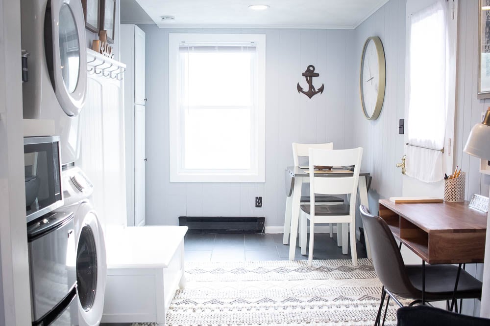 A washing machine and dryer are positioned side by side against a wall. A small dining table with white chairs is situated near a window, while a desk and chair are also present in the space. Light-colored walls and a nautical-themed clock add to the room's design.