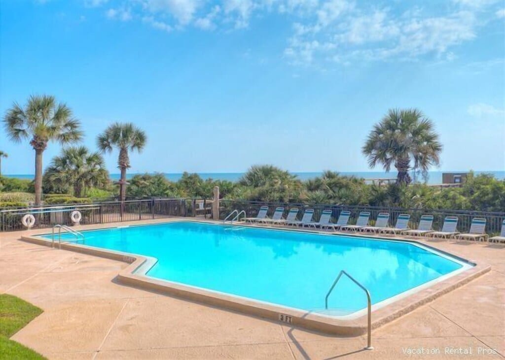 A heated pool is surrounded by lounge chairs, with palm trees and lush greenery in the background. The ocean is visible in the distance, under a clear blue sky, creating a refreshing environment for relaxation.