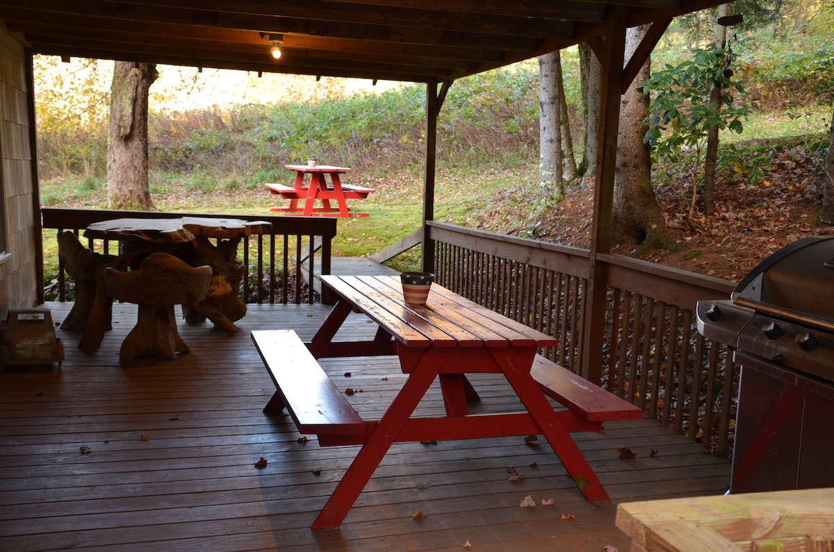 An outdoor patio area is presented, featuring a red picnic table and a wooden dining table with animal-shaped benches. A grill is positioned nearby, and natural surroundings are visible in the background, with trees and greenery enhancing the peaceful setting.