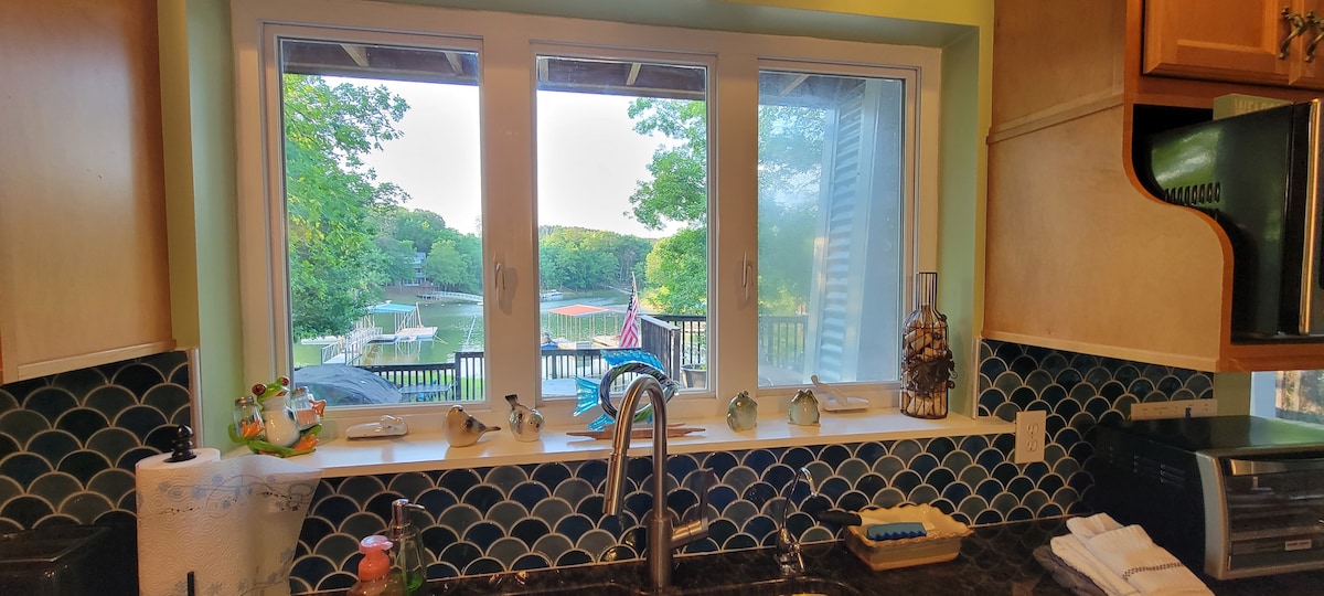 A kitchen window offers a view of the serene lakeside, framed by large panes. The countertop features a modern faucet, with various decorative items positioned below the window, including small figurines. The vibrant green trees reflect the calm water outside.