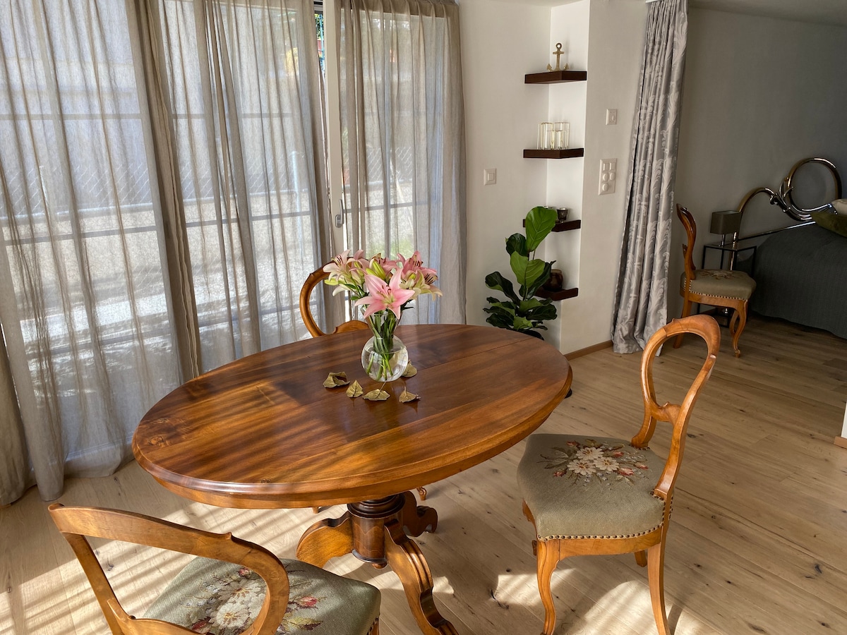A round wooden dining table is positioned near floor-to-ceiling windows, adorned with a vase of fresh flowers. Two upholstered chairs, featuring floral patterns, complement the table. Soft natural light filters through sheer curtains, enhancing the inviting dining area.