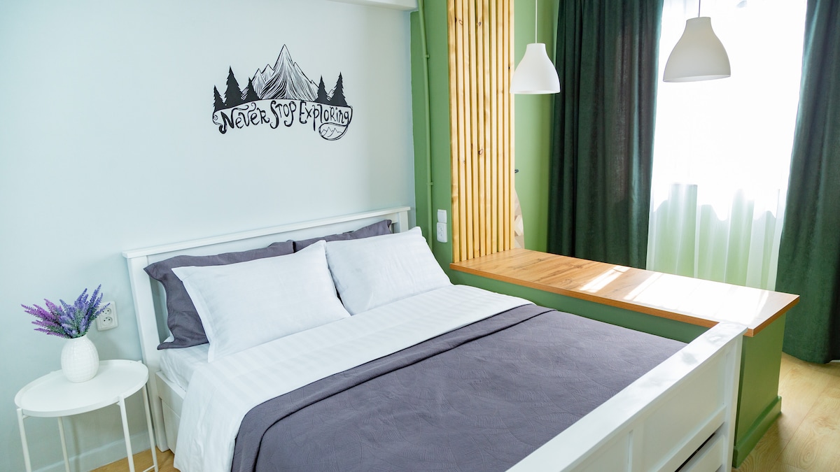 A serene sleeping area is highlighted by soft white bedding on a spacious bed. Natural light filters through sheer green curtains, complementing the muted walls. A wooden shelf is positioned behind the bed, and a small round table holds fresh lavender, contributing to a calm atmosphere.