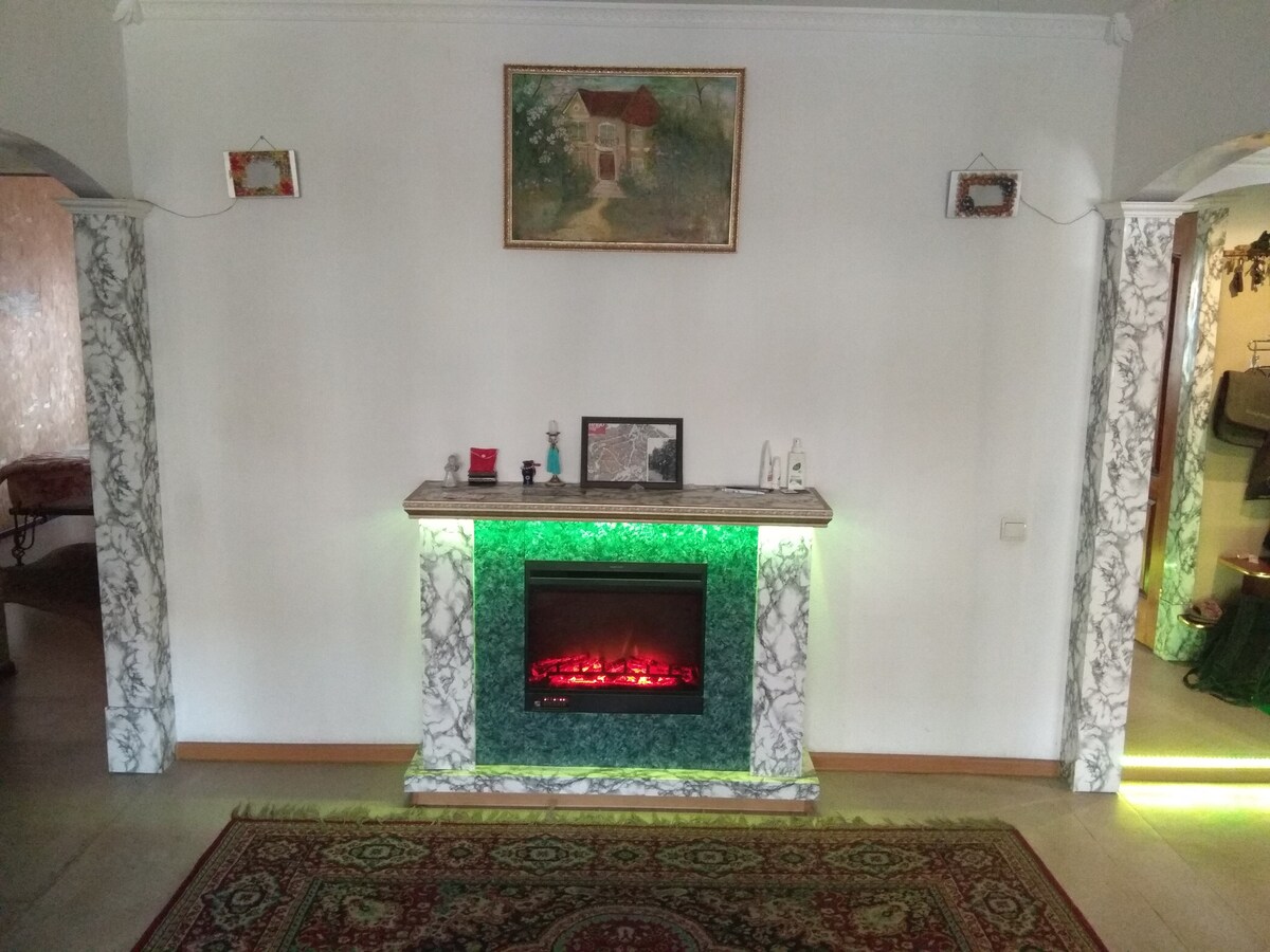 A decorative fireplace with a green glowing base is set against a light-colored wall. A framed artwork is displayed above, adding an artistic touch. The marble columns on either side feature intricate designs, complemented by a patterned area rug on the floor.