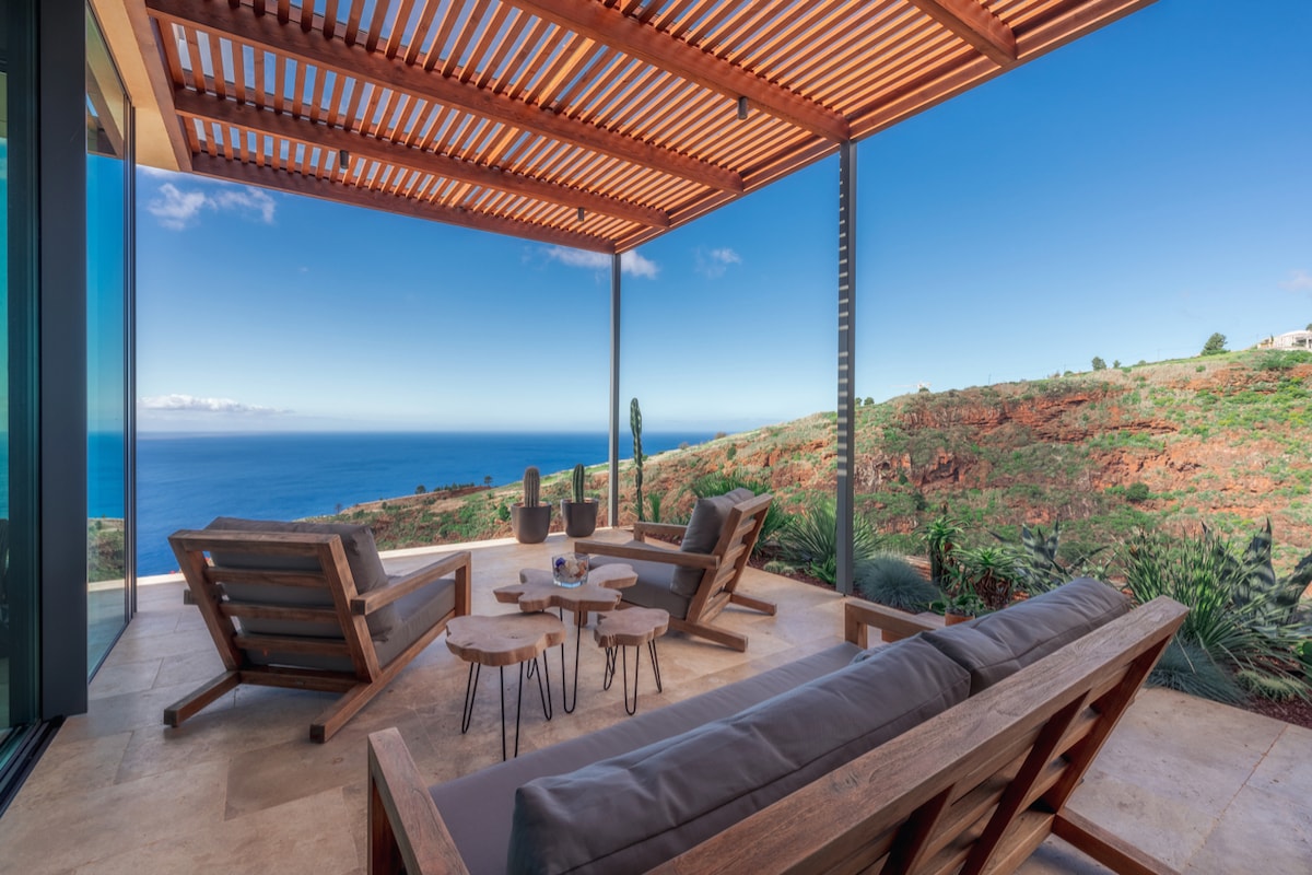 An outdoor seating area features comfortable wooden lounge chairs and matching side tables. The space provides expansive views of the Atlantic Ocean and the surrounding cliff landscape, framed by large glass panels and a wood slatted roof that offers shade.