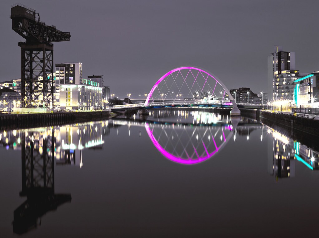 A nighttime view of the Clyde River is visible, featuring a vibrant pink arch bridge reflecting on the water's surface. Nearby, a distinctive structure stands tall alongside illuminated buildings, creating a harmonious cityscape.