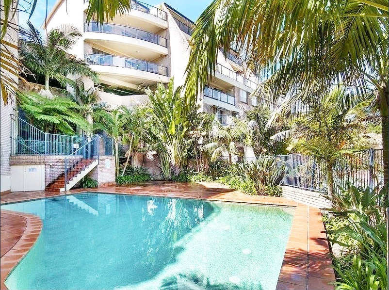 A serene pool area is framed by lush tropical vegetation and palm trees. The clear water reflects sunlight, creating a calming atmosphere. Surrounding the pool, spacious terraces with stone pathways provide access to the apartment complex, enhancing the outdoor experience.