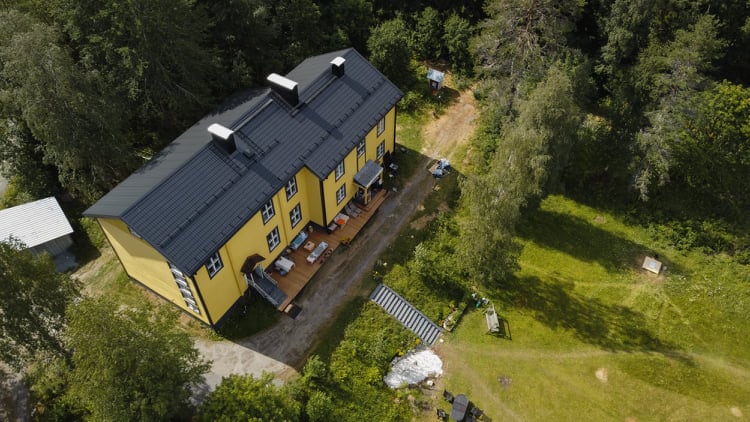 An aerial view captures the two-story, yellow villa surrounded by lush greenery. A spacious deck is visible at the back, furnished with outdoor seating. The pathway leads to an expansive yard, showcasing natural surroundings and additional outdoor elements.