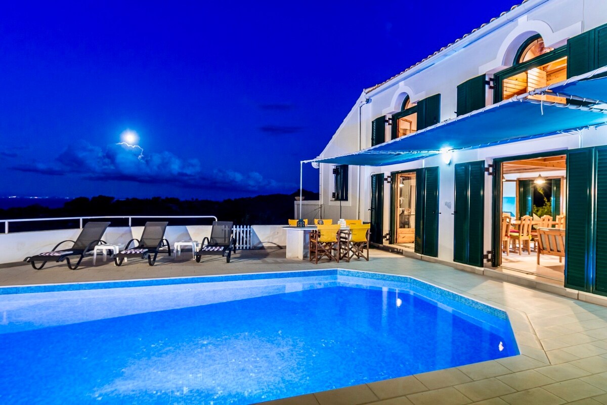 A spacious terrace surrounds a private swimming pool, illuminated by soft lighting under the night sky. Sunbeds are arranged alongside the pool, with dining chairs visible near the open doors leading to an inviting indoor space.