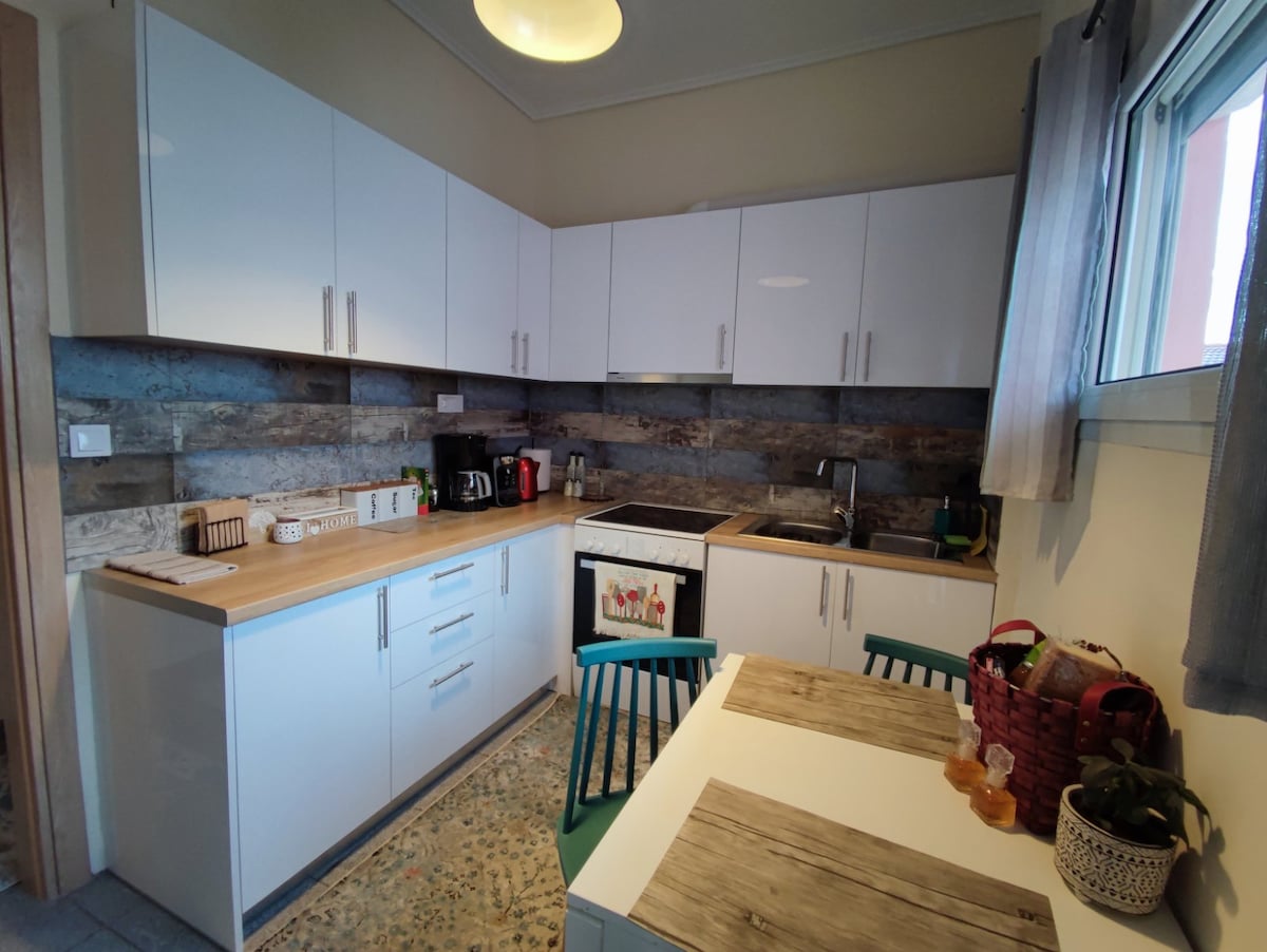 The kitchen features modern cabinetry in a clean white finish, complemented by wooden accents on the walls. A compact dining table with teal chairs is positioned neatly under a window, allowing natural light to brighten the space. Essential appliances are arranged for convenience.