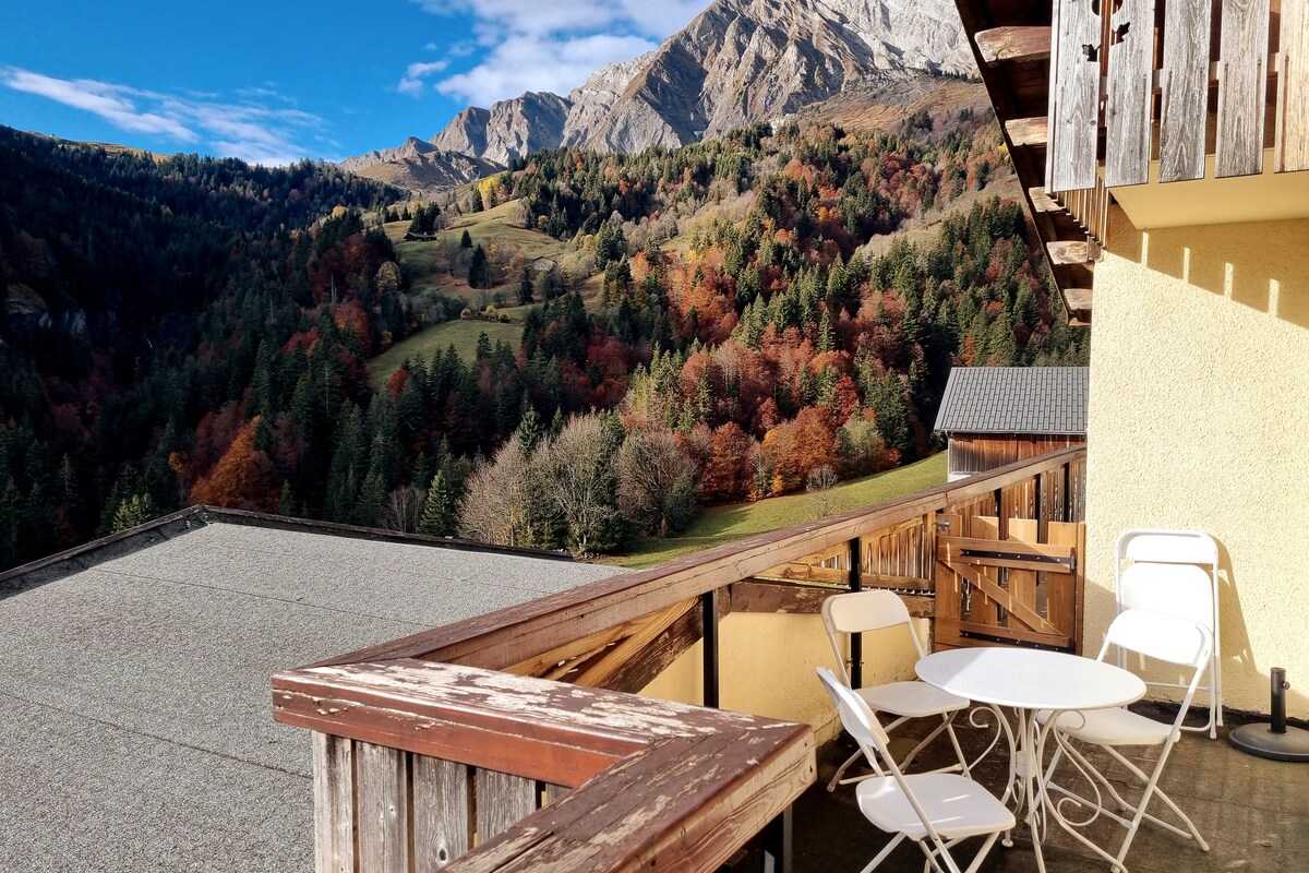 A spacious terrace is presented, featuring a round table and four chairs, all set against a backdrop of vibrant fall colors in the surrounding mountains. The landscape showcases a blend of greens and oranges, with distant peaks framing the view.