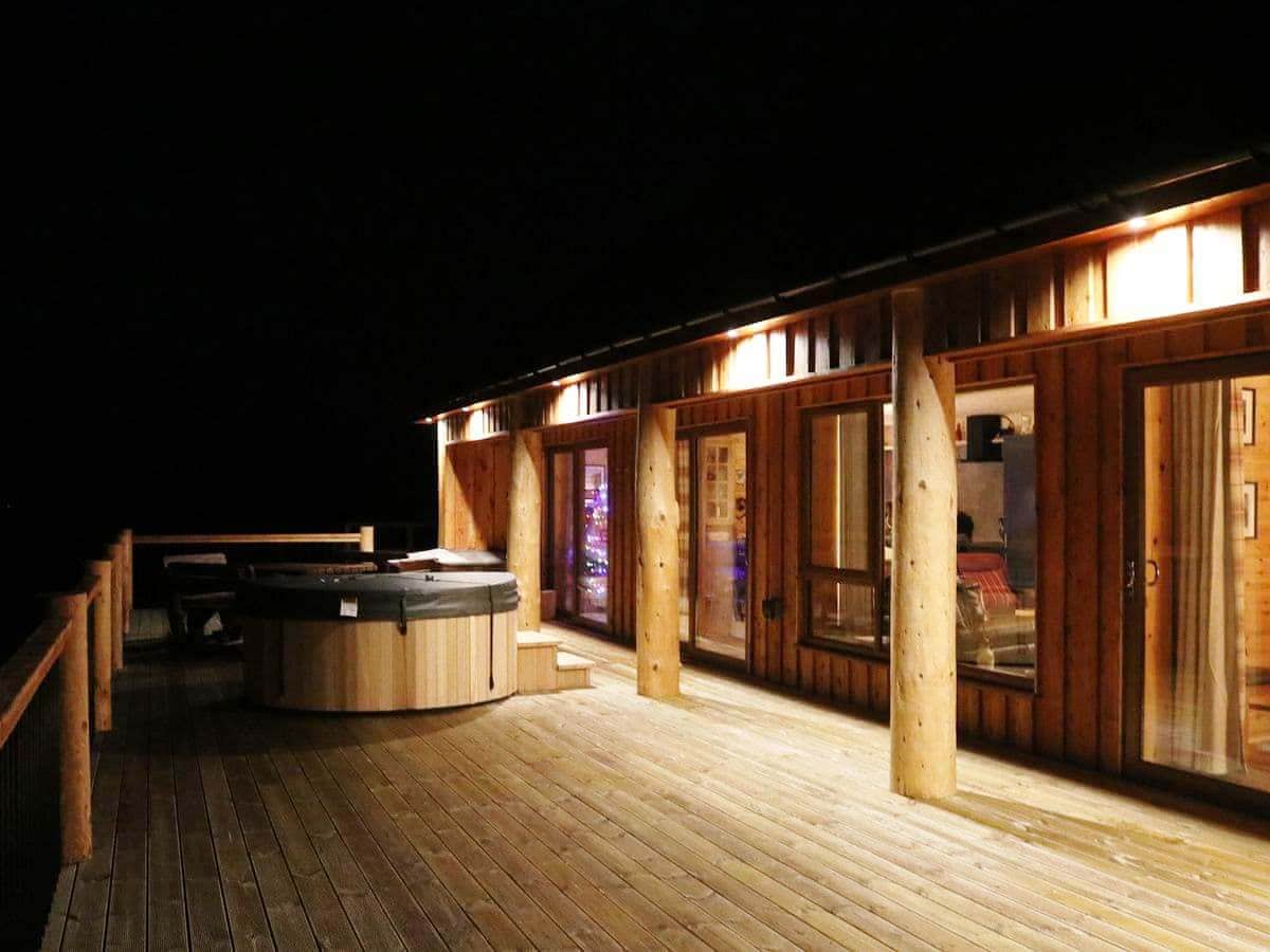A spacious wooden deck is illuminated softly at night, featuring a private hot tub adjacent to large glass doors that open into the lodge. The natural wood accents of the structure are highlighted, enhancing the tranquil outdoor setting.