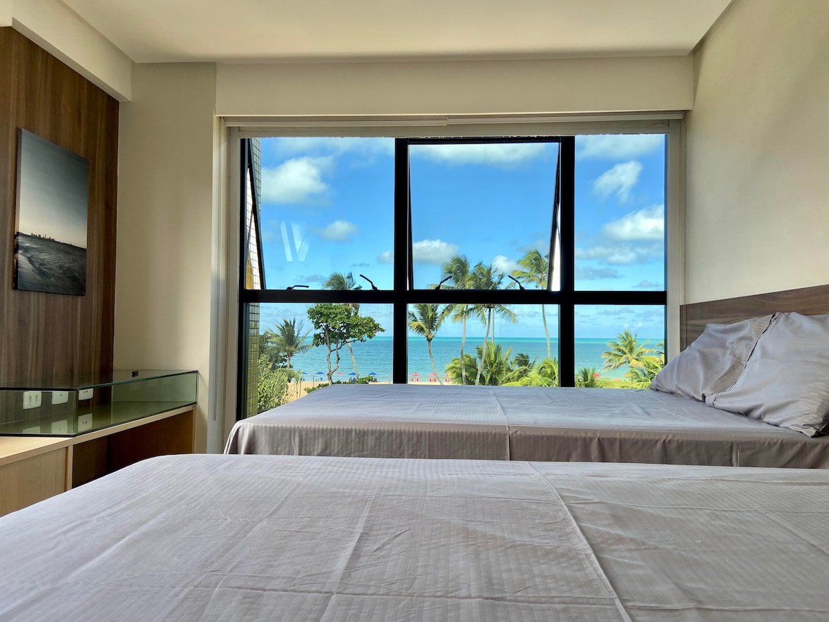 The bedroom features two spacious beds positioned in front of large windows, offering a direct view of the ocean and palm trees. Natural light fills the room, highlighting the minimalist decor and soft bedding.