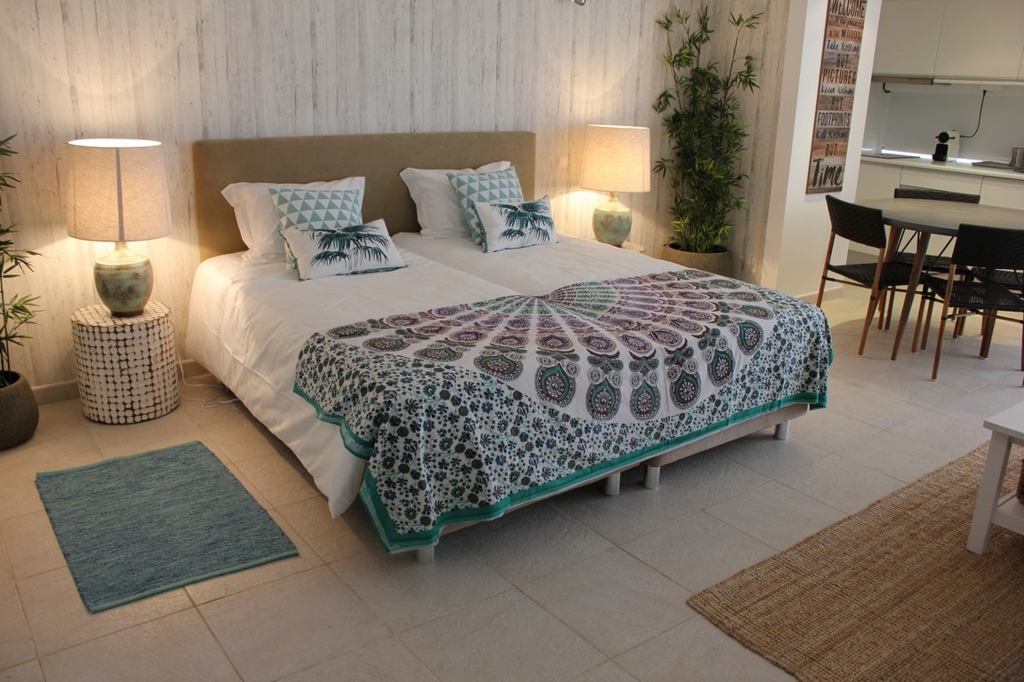 A spacious bedroom features two beds with colorful patterned covers. Warm light from lamps beside each bed creates a soft ambiance. A dining area with a small table and chairs is visible in the background, complemented by elements of nature, such as potted plants.