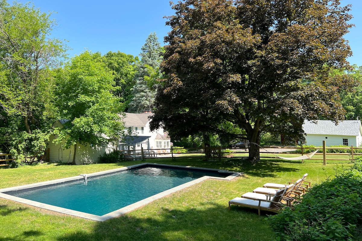 A heated seasonal pool is centered within a lush green lawn, surrounded by mature trees. Comfortable lounge chairs line the pool's edge, offering a spot for relaxation. The property’s charming farmhouse and additional buildings can be seen in the background, nestled among the greenery.