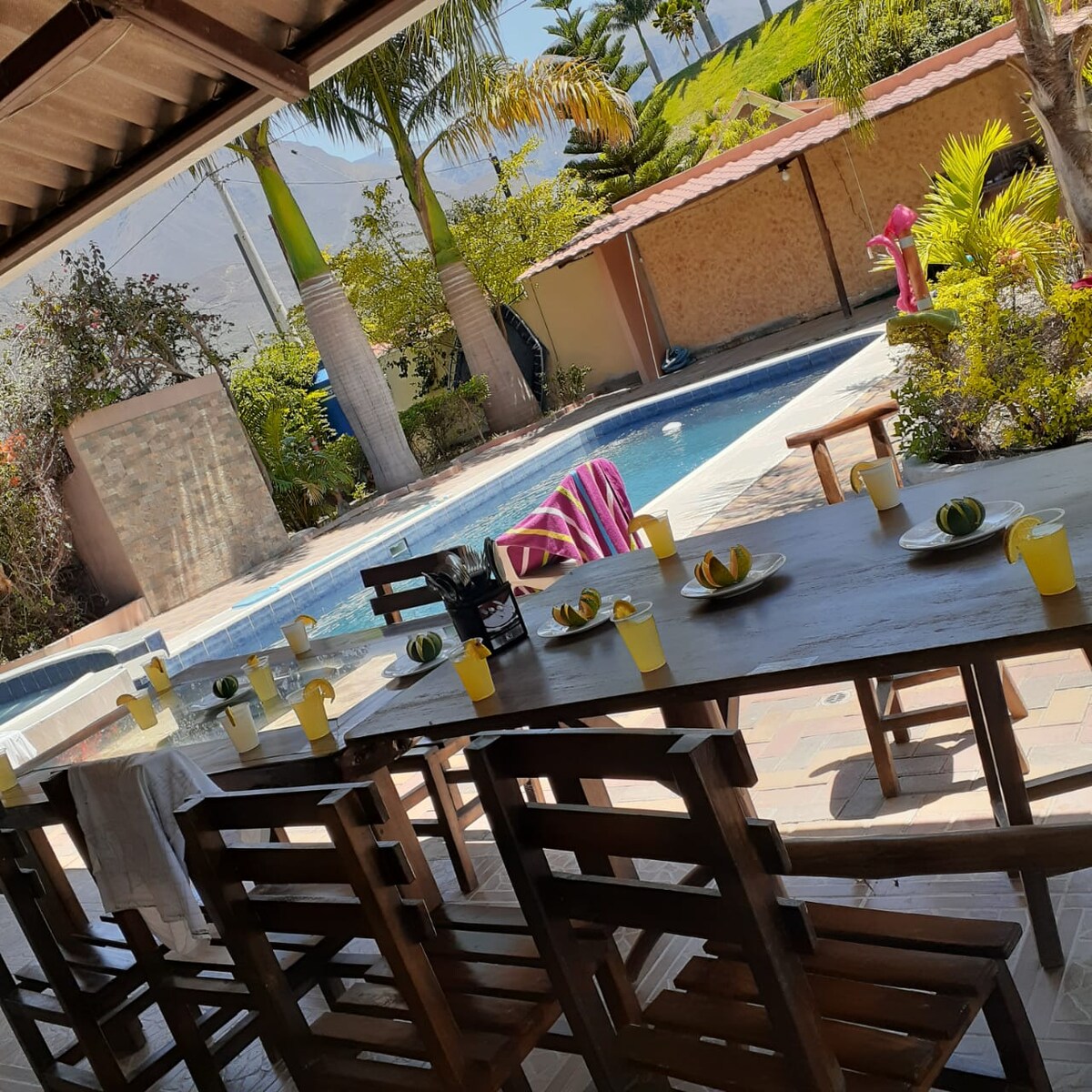A spacious outdoor dining area features a long wooden table set with yellow cups and garnished with limes. A sunlit pool is visible in the background, surrounded by tropical plants, lounge chairs, and a vibrant beach towel. The setting offers a relaxed atmosphere for gatherings.