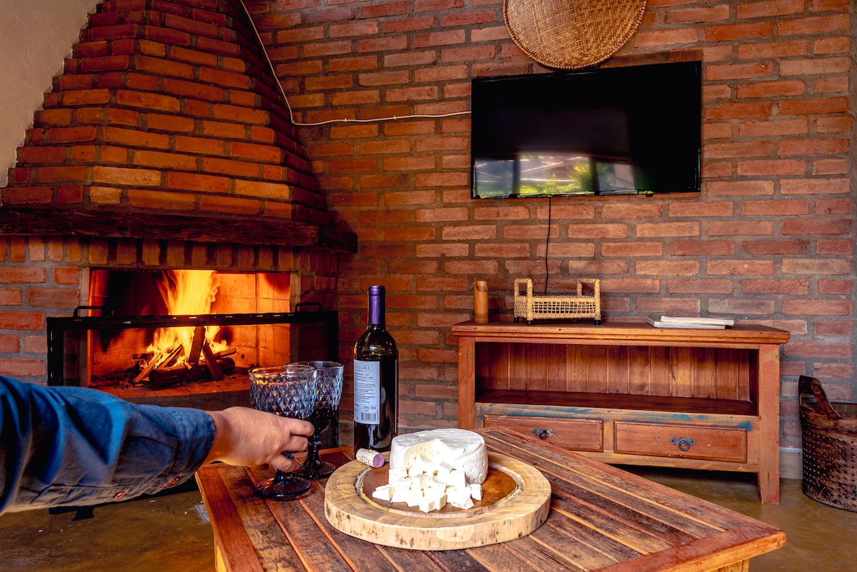 An internal space features a brick wall, enhancing the cozy atmosphere. A fireplace provides warmth with a gentle flame. A wooden table displays a cheese platter and a bottle of wine beside a glass. An entertainment area includes a mounted television, completing the inviting setup.