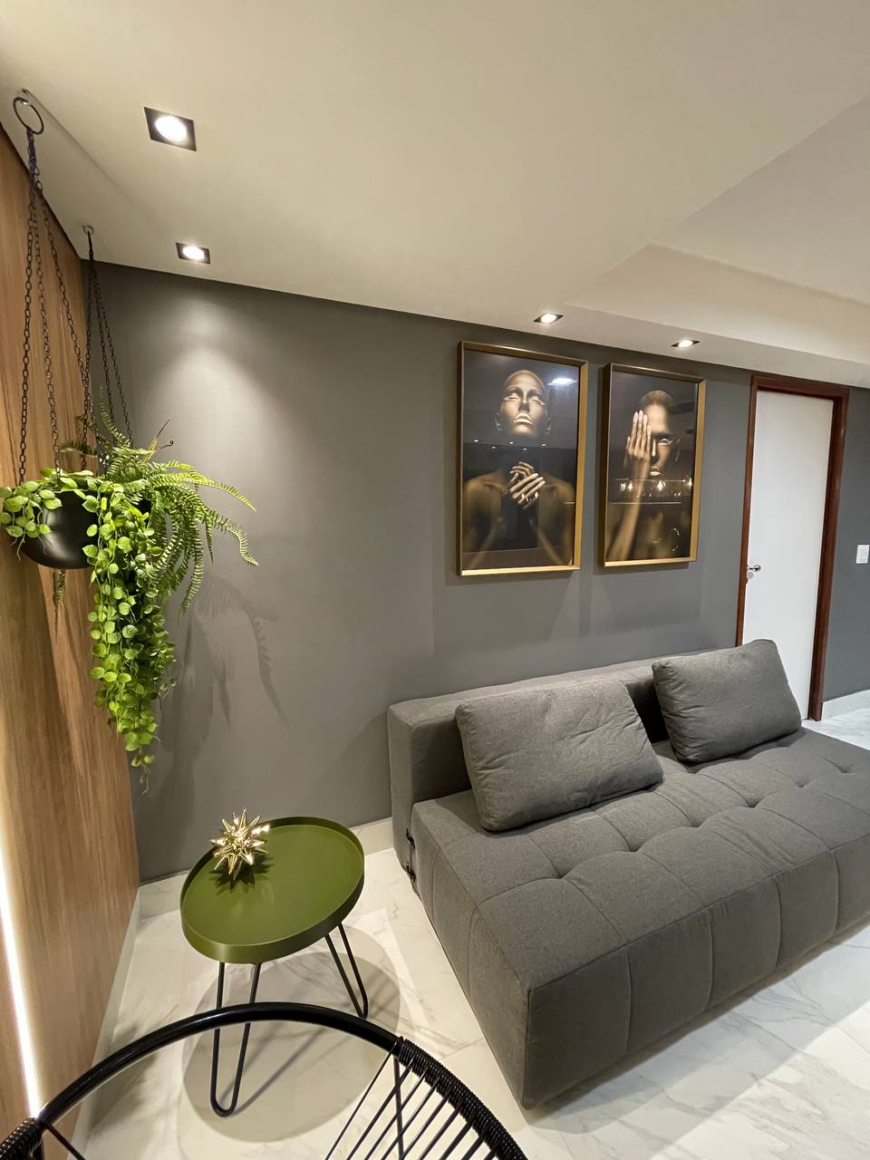 A modern living space is depicted, featuring a gray sofa and a small green side table. Two framed artworks hang on the wall, complemented by a hanging plant to the left. Soft lighting from recessed fixtures creates a warm ambiance.