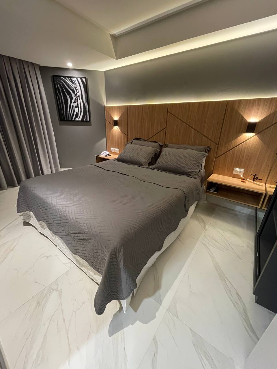 A comfortable sleeping area is featured with a spacious bed covered in a grey quilt. Wooden accents and bedside lamps create a subtle ambiance. Light-colored curtains frame the window, while a piece of wall art adds a touch of design to the modern room.
