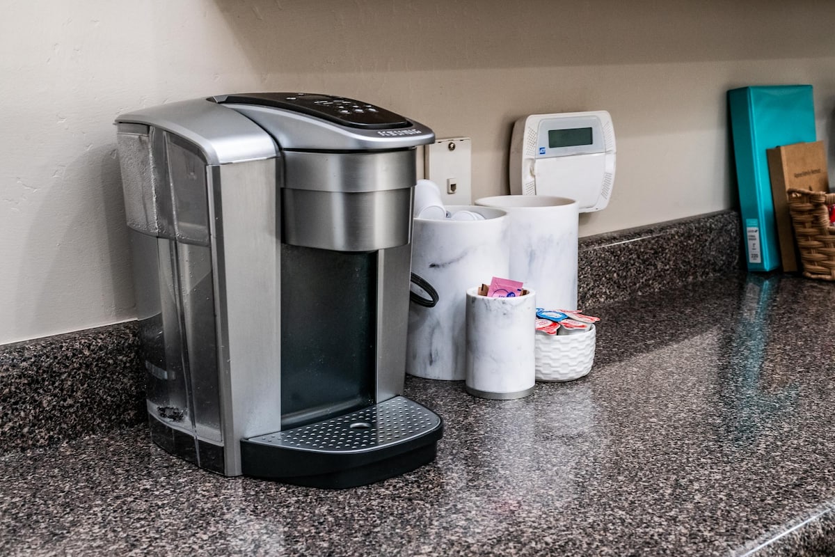A coffee maker is positioned on a granite countertop, accompanied by marble canisters that store assorted coffee supplies. Nearby, a programmable thermostat and a basket containing miscellaneous items are also visible, creating a functional kitchen area.