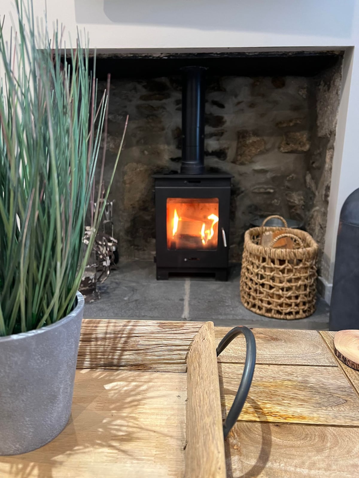 A wood burning stove is positioned against a stone wall, radiating warmth. In the foreground, a textured pot containing green grass-like plants sits on a rustic wooden table, with a woven basket nearby, adding to the cozy ambiance of the space.