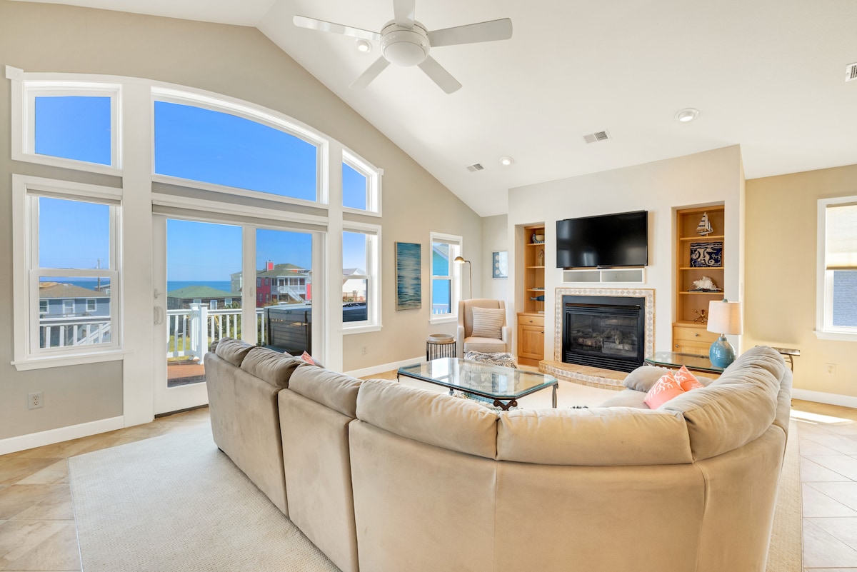 A spacious living area features large windows showcasing ocean views and allowing natural light to fill the room. A comfortable sectional sofa surrounds a glass coffee table, with a TV and fireplace positioned against the wall. The atmosphere is bright and inviting.