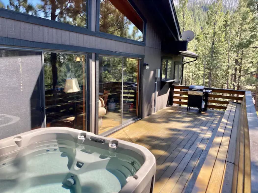 A spacious deck features a hot jacuzzi and a grill, surrounded by pine trees. Large glass doors provide a view into the living area, where natural light enhances the inviting atmosphere. The wooden deck offers ample space for relaxation and enjoyment of the outdoors.