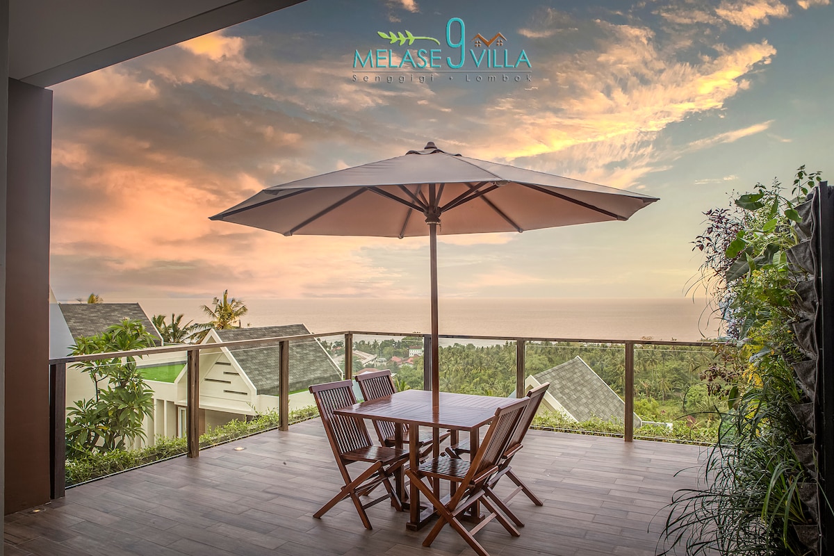 An outdoor terrace is furnished with a wooden dining table and four chairs, shaded by a large umbrella. The view includes lush greenery and rooftops, with the ocean visible in the distance under a colorful sky.