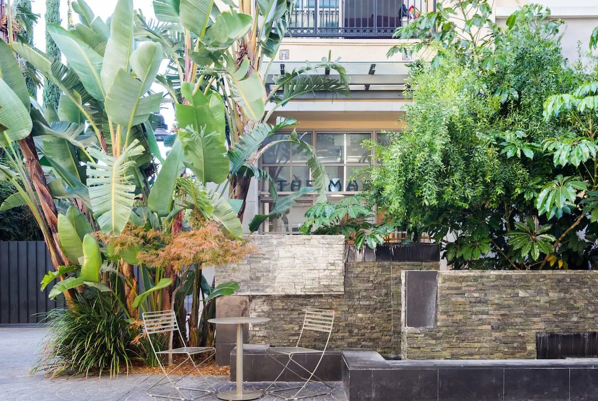 Lush greenery surrounds a tranquil outdoor space featuring a small water feature. Two metal chairs and a circular table are positioned nearby, creating an inviting spot for relaxation amidst the vibrant foliage.
