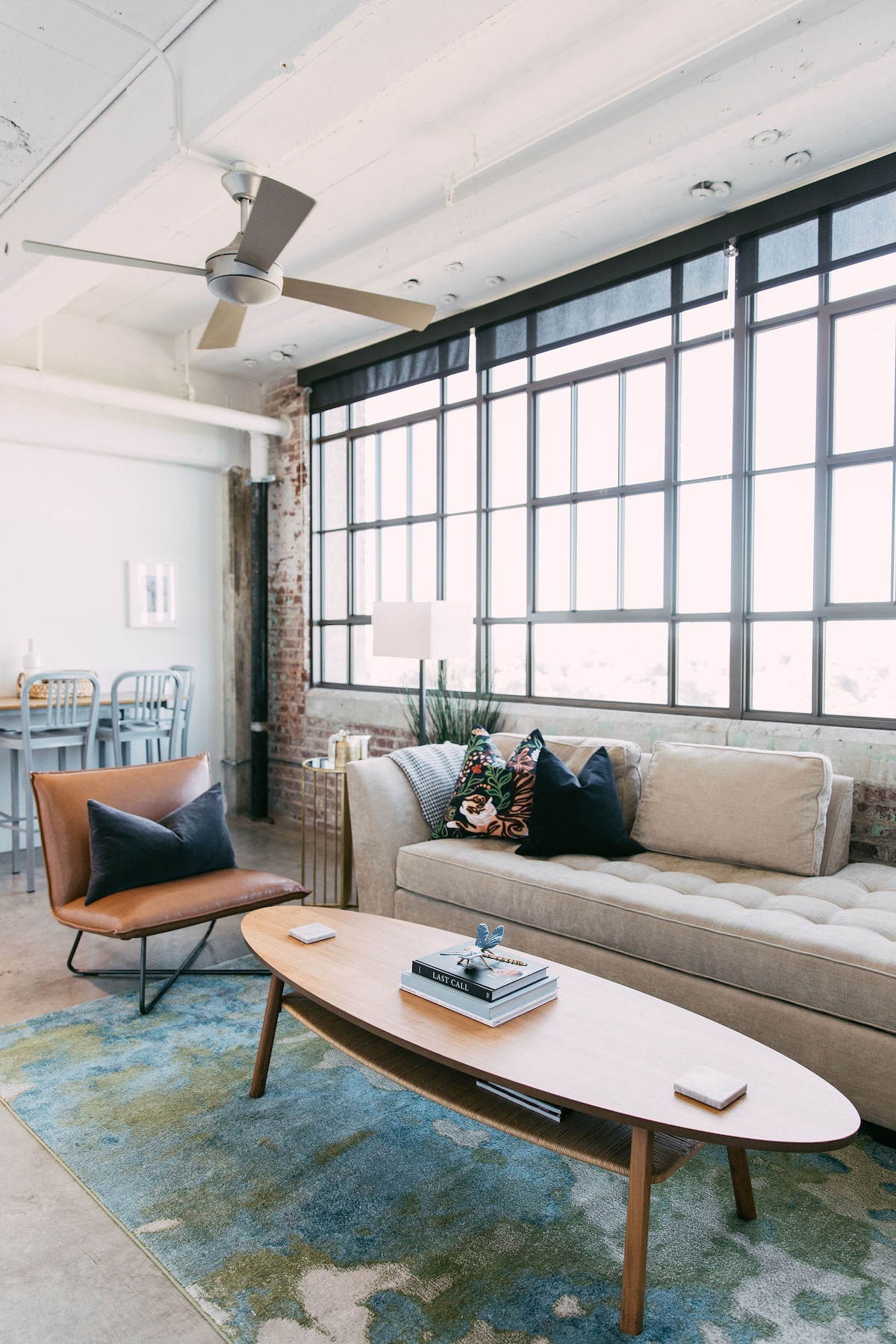A spacious living area features a light-colored sofa adorned with patterned cushions. A wooden coffee table sits atop an area rug, accompanied by a stylish chair. Large industrial windows allow ample natural light, while exposed brick adds character to the room.