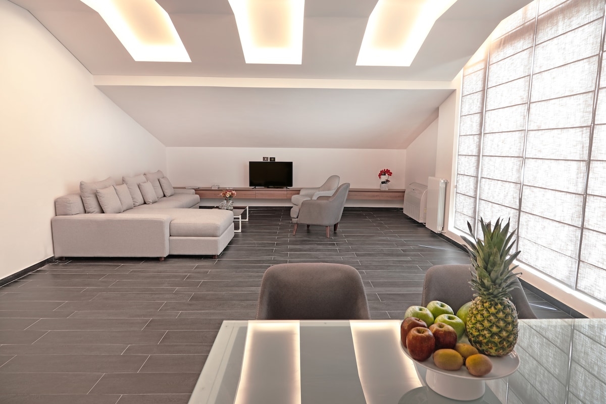A modern living area is furnished with a spacious gray sectional sofa and two lounge chairs. A sleek entertainment unit displays a television. Natural light filters through a large window, and a glass dining table features a bowl of fresh fruit, including apples and pineapple.