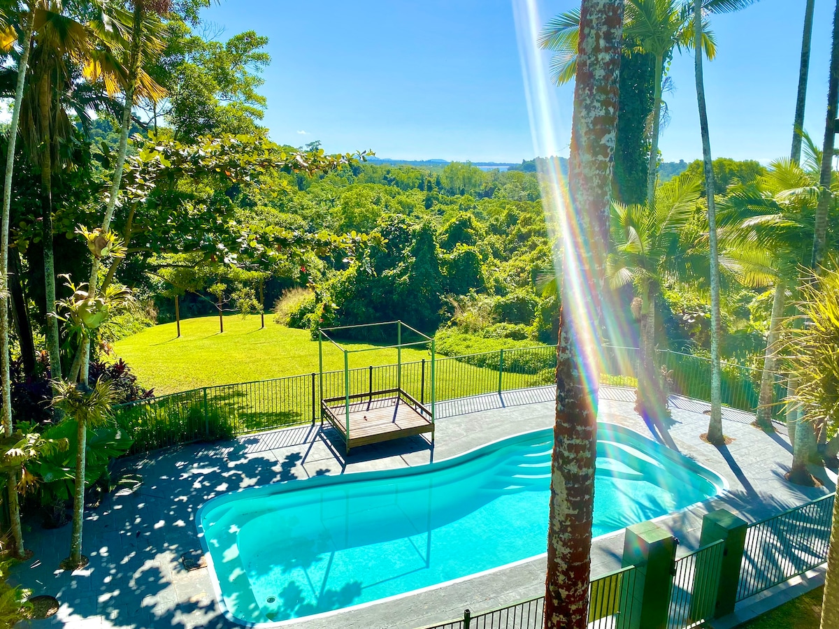 A serene area features a sparkling swimming pool surrounded by lush greenery. A wooden deck provides a space for relaxation, and expansive views of the vibrant landscape can be seen in the distance under clear blue skies.
