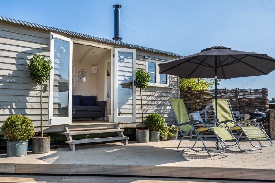A charming patio area is presented with two reclining chairs and an umbrella, inviting relaxation. The entrance of the hut, framed by planters, showcases double doors that open to the living space, complete with a comfortable sofa and wood-burning stove.