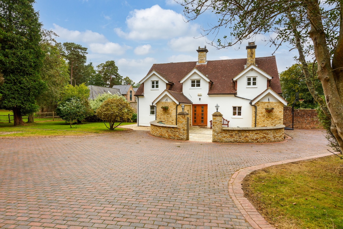 A well-maintained cottage is surrounded by lush greenery and a spacious driveway. The home features a charming facade with a combination of white walls and stone accents, illuminated by soft natural light. A decorative stone wall borders the entrance, enhancing the welcoming feel.
