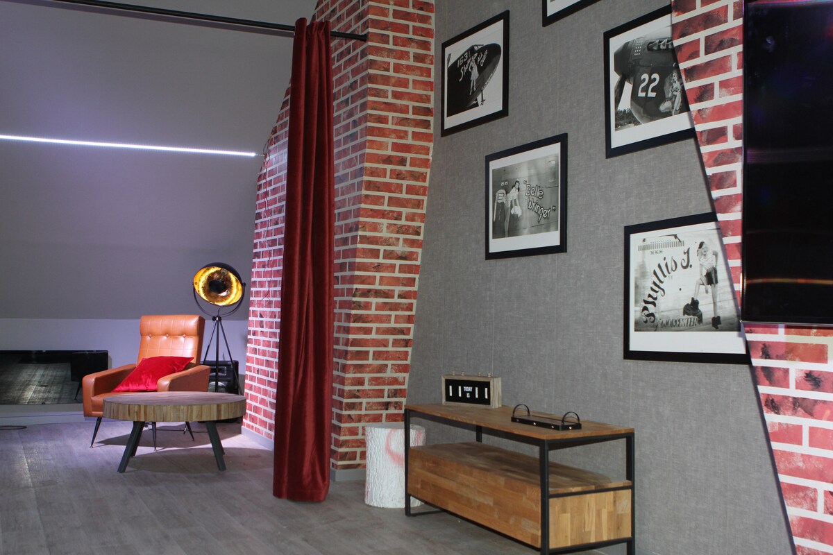 A modern living space is featured, showcasing a mix of textured brick and fabric wall finishes. A stylish orange chair provides a pop of color, while framed black-and-white photographs are displayed on the wall. A wooden coffee table and decorative items add functionality to the area.