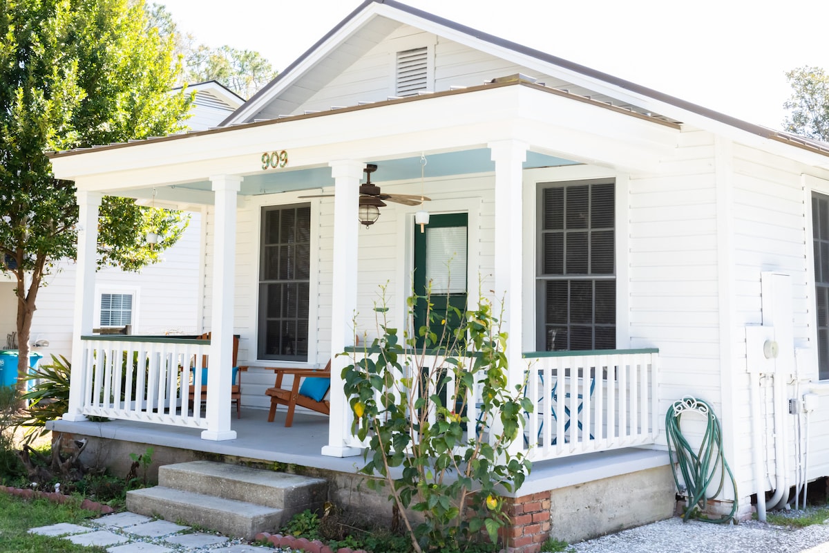 The front porch is framed by white columns and a light green door. Blue seating is arranged on the porch, welcoming guests to relax. A small garden featuring greenery complements the home's exterior, which is painted in crisp white.