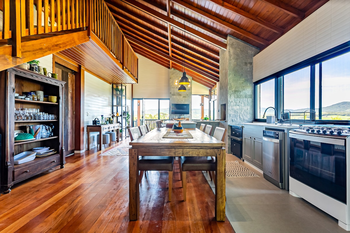 A spacious kitchen and dining area features high, exposed wooden ceilings and large windows, providing ample natural light. A long wooden table is surrounded by chairs, while a modern kitchen boasts stainless steel appliances and a rustic wooden cabinet filled with glassware.