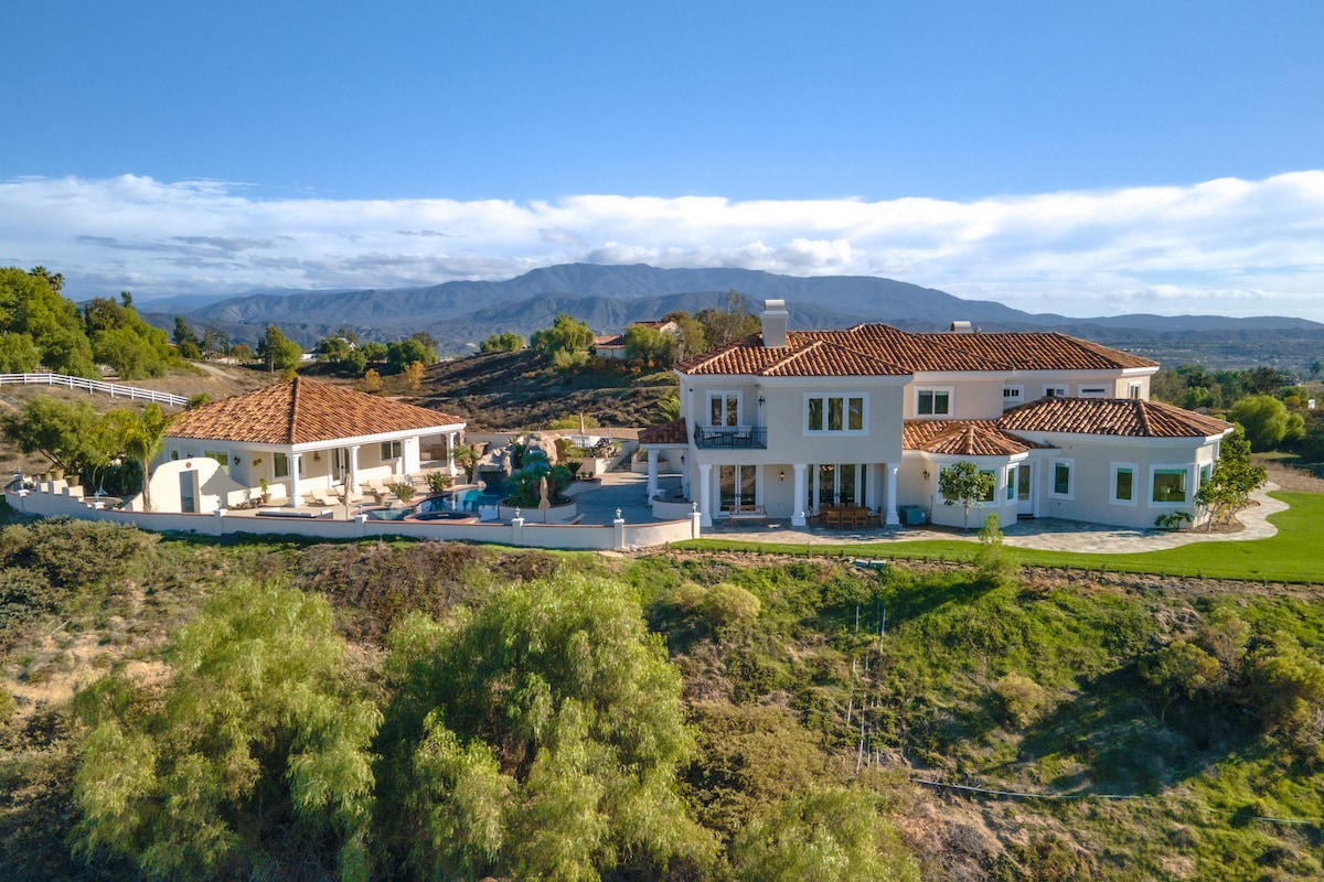 The estate is showcased from an aerial view, highlighting its two-story design and terracotta roof. Lush greenery surrounds the property, while the stunning Temecula Valley mountains create a serene backdrop. The outdoor space features a pool area, emphasizing the property’s impressive location.