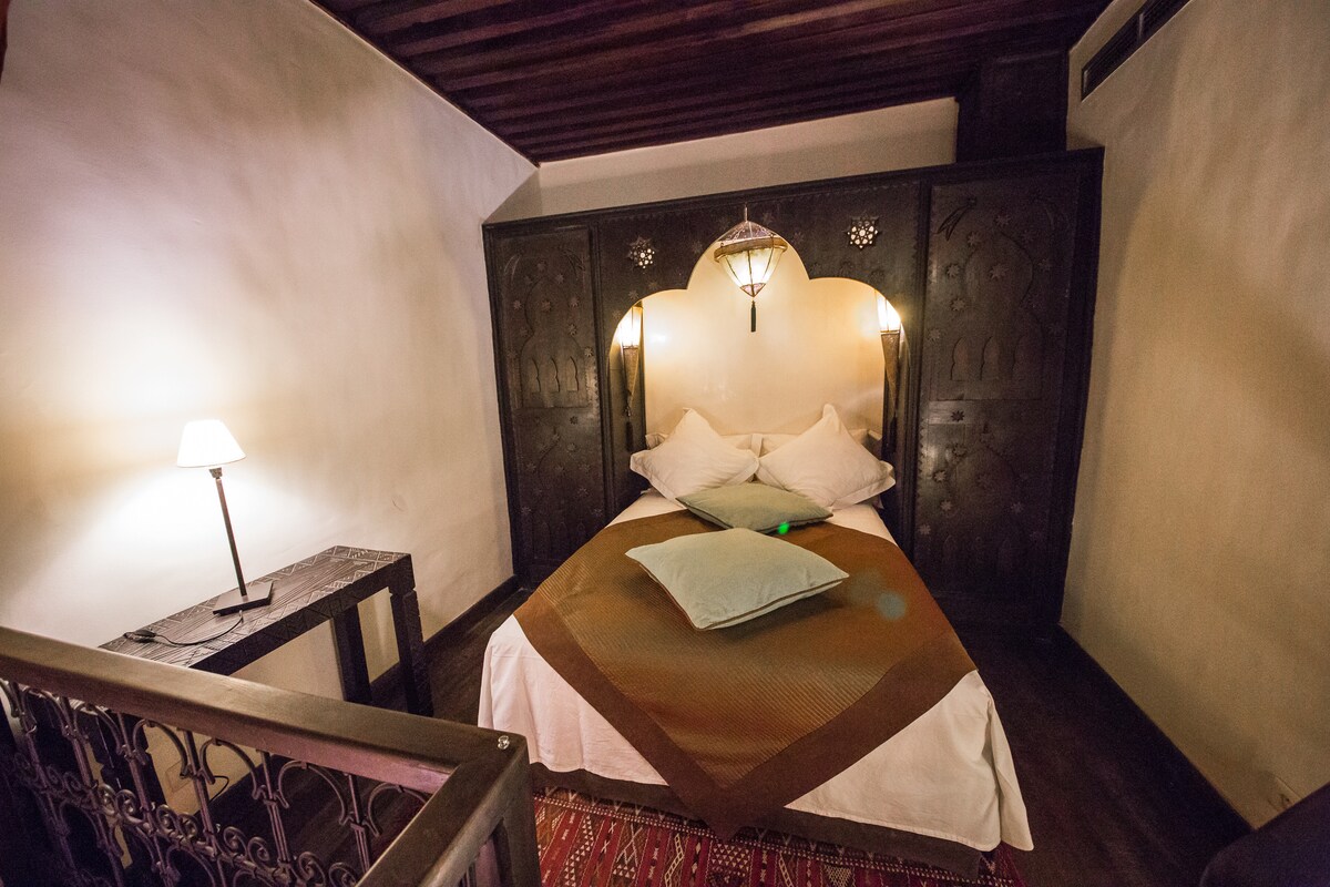 A sleeping area features a spacious double bed with light-colored linens, placed against a decorative wooden headboard. Soft lighting is provided by bedside lamps, and a traditional carpet adds warmth to the wooden floor. An inviting atmosphere is created by the room's unique design.