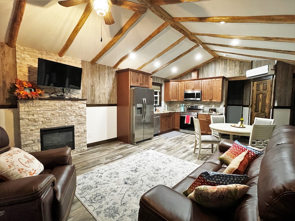 A cozy living space combines rustic elements with modern amenities. A stone fireplace is paired with a large flat-screen TV. The kitchen features stainless steel appliances and wood cabinetry, while a dining table and comfortable seating area contribute to a welcoming ambiance.