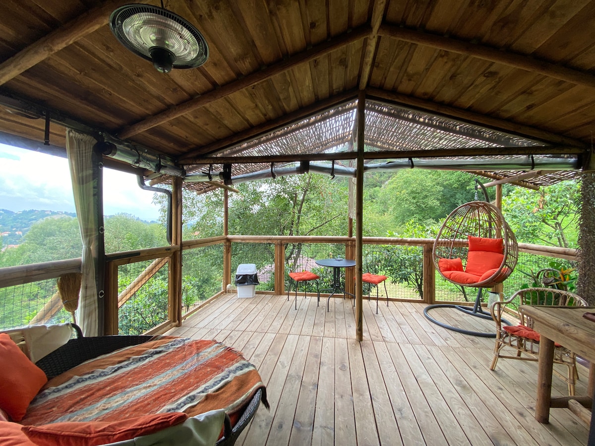 An open terrace features wooden beams and a thatched roof, providing a shaded seating area. A circular hanging chair and a traditional table are positioned among the greenery, while a plush, round lounge chair invites relaxation. Scenic views of the surrounding landscape can be observed.