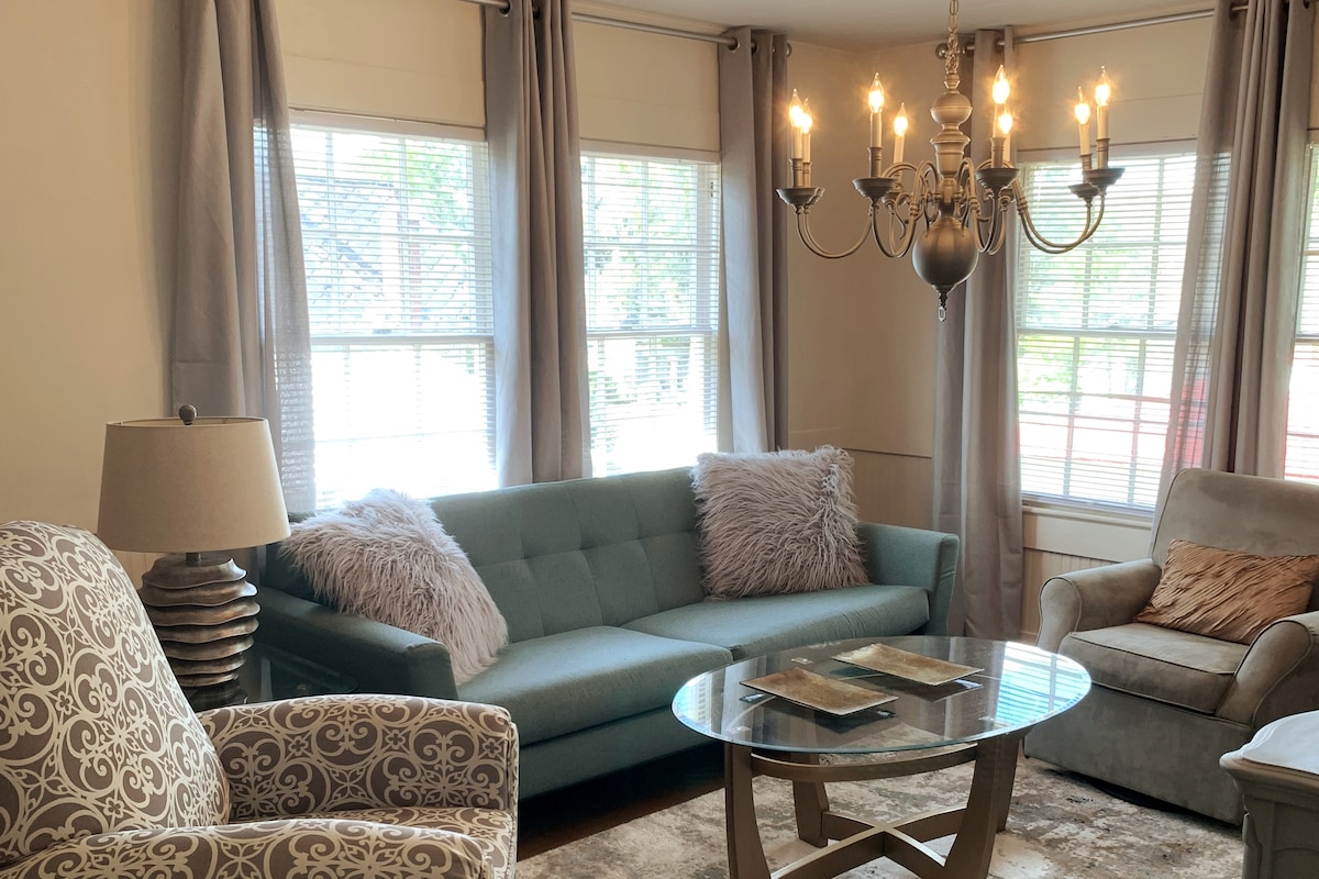 A bright living area features large windows framed by light gray curtains, allowing natural light to fill the space. A soft blue couch with fluffy pillows is complemented by a round glass coffee table, and a stylish chandelier provides overhead lighting.